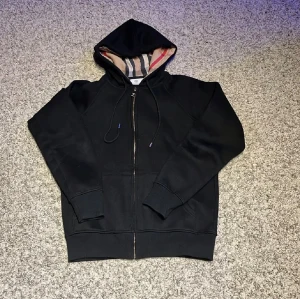 Svart Burberry hoodie med dragkedja - Svart hoodie från Burberry med klassisk dragkedja och huva. Insidan av huvan har Burberrys ikoniska rutiga mönster i beige, svart och rött. (Den bruna lappen tillkommer inte)