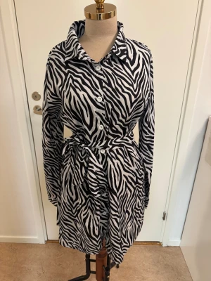 Zebramönstrad skjortklänning från SHEIN - Snygg zebramönstrad skjortklänning från SHEIN i svart och vitt. Klänningen har långa ärmar, knappar framtill och ett matchande knytbälte i midjan. Perfekt för dig som vill sticka ut med ett djurmönster och gillar en relaxed men trendig look.