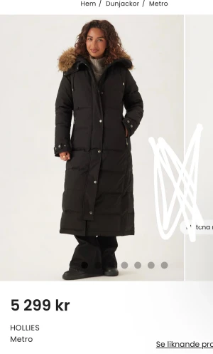 Svart lång dunjacka Hollies Metro - Lång svart dunjacka från Hollies Metro med stor huva och fluffig ÄKTA avtagbar päls. Jackan har dragkedja och knappar framtill, två sidofickor med dragkedja och quiltad design. Perfekt för kalla dagar och riktigt snygg siluett. jätte varm i helt nytt skick
