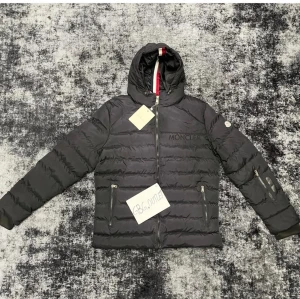 Svart Moncler pufferjacka med huva - Svart pufferjacka från Moncler med huva som har en snygg röd och vit rand på insidan. Jackan har broderad Moncler-logga på bröstet, dragkedjor på fickorna och ärmarna samt quiltad design. Perfekt för kalla dagar och riktigt stilren look.
