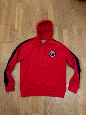 Röd hoodie från FILA med blå detaljer - Snygg röd hoodie från FILA i storlek M med blå ränder längs ärmarna och ett broderat FILA-märke på bröstet. Tröjan har huva med snörning och är tillverkad i mjuk bomull. Perfekt för dig som gillar sportig och avslappnad stil.