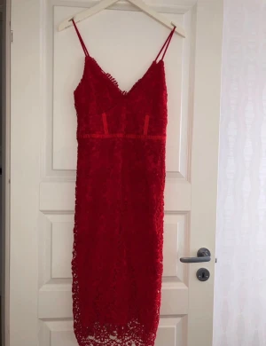 Röd spetsklänning med smala axelband - Röd spetsklänning med smala axelband och v-ringad hals. Klänningen har en figurnära passform och vacker spetsdetalj över hela plagget. Perfekt längd som slutar under knäet och fina detaljer vid bysten. Snygg och trendig look!