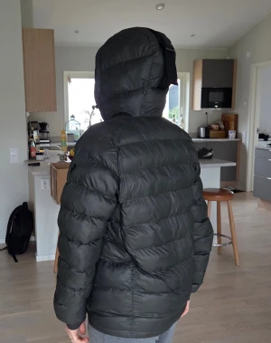 Mörkgrön  dunjacka från Peak Performance - Mörkgrön dunjacka från Peak Performance med huva och dragkedja framtill. Jackan har quiltad design, ribbade muddar vid ärmslut och en snygg rem på huvan. Perfekt för kalla dagar och har en clean, sportig look.