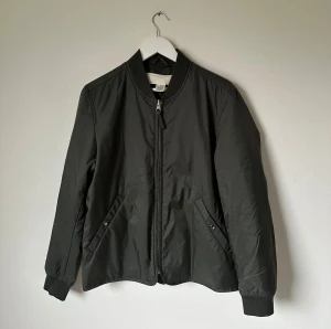 Mörkgrön bomberjacka från ARKET - Snygg mörkgrön bomberjacka från ARKET med klassisk ribbad krage och muddar. Jackan har dragkedja framtill, två snedställda fickor med knapp och är tillverkad i polyester. Perfekt för dig som gillar stilren och enkel design. 5N14