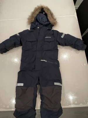 Mörkblå Didriksons vinteroverall - Säljer en mörkblå Didriksons vinteroverall med huva och fuskpäls, flera fickor och reflexdetaljer. Overallen har dragkedja och knappar framtill samt förstärkta partier på knäna. Perfekt för kalla dagar och snölek!