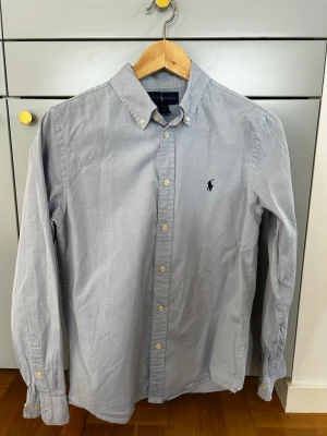Ljusgrå skjorta från Ralph Lauren S - Klassisk ljusgrå skjorta från Ralph Lauren med button-down krage och broderad logga på bröstet. Skjortan har långa ärmar, vita knappar och är tillverkad i mjuk bomull. Perfekt för en clean och stilren look. Storlek S