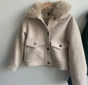 Beige teddyjacka med fluffig krage - Säljer en beige teddyjacka från H&M med fluffig krage i fuskpäls. Jackan har två stora fickor med knappar framtill och stängs med knappar. Croppad modell som ger en cool och trendig look, perfekt för höst och vinter.