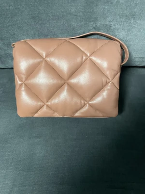 Quiltad brun axelväska från Stand Studio - Snygg quiltad axelväska i brun skinnimitation från Stand Studio. Väskan har en mjuk, puffig look med diamantmönster, guldfärgade detaljer och dragkedja på insidan. Perfekt storlek för att få plats med allt du behöver och har en stilren design.