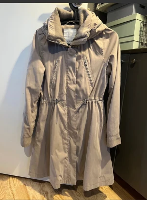 Beige parkajacka från Elvine XS - Snygg beige parkajacka från Elvine med dragkedja och tryckknappar fram. Jackan har två fickor med dragkedja upptill samt två fickor nedtill och dragsko i midjan. Jackan har en huva som kan rullas in i kragen. Mjuk skön krage. Längd ca 85cm.