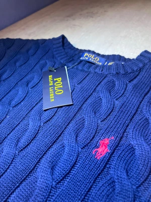 Marinblå kabelstickad tröja Polo Ralph Lauren - Snygg marinblå kabelstickad tröja från Polo Ralph Lauren med klassisk rund hals och långärmad design. Tröjan har det ikoniska rosa broderade logot på bröstet och ribbade muddar vid ärmslut och nederkant. Perfekt för dig som gillar stilren och tidlös look.