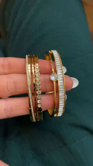 Guldiga armband med strassdetaljer - Säljer ett set med fyra guldiga armband i metall, dekorerade med glittrande strass och kristaller. Ett armband har en stor rund kristall i mitten, medan de andra har olika mönster av små stenar och geometriska former. ROSTFRITT💛.