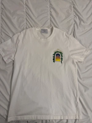 Vit t-shirt från Casablanca med tryck - Vit t-shirt från Casablanca med färgstarkt tryck av en byggnad och gröna blad på bröstet och ryggen. Rund hals och korta ärmar. Materialet är mjuk bomull och trycket har detaljer i gult, blått, grönt och rött. Perfekt för dig som gillar unika prints och streetwear.