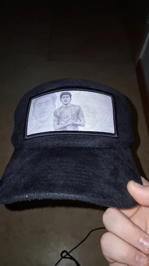 Svart truckerkeps med tryck - Säljer en svart truckerkeps med mesh på sidorna och baksidan. Framtill finns en rektangulär patch med ett svartvitt tryck av en person. Kepsen har böjd skärm och justerbar storlek. Priset är ej hugget i sten