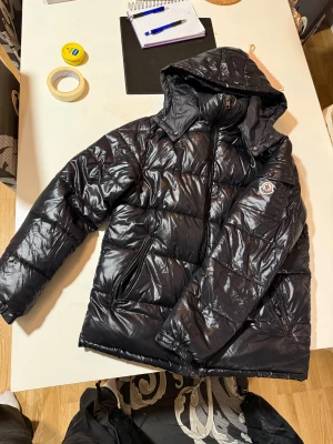 Svart Moncler pufferjacka med huva - Svart pufferjacka från Moncler med glansig finish, huva och dragkedja framtill. Jackan har två sidofickor med dragkedja och en ficka på ärmen med Moncler-logga. Insidan har en unik serietidningsetikett. Perfekt för kalla dagar och riktigt snygg streetstil. Pris är diskuteras 