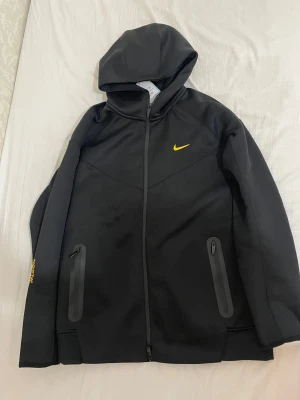 Nike NOCTA svart tracksuit set - Säljer ett svart tracksuit set från Nike NOCTA med gul logga och detaljer. Hoodien har dragkedja, huva och två fickor med dragkedja. Byxorna har snörning i midjan, dragkedjeficka och NOCTA-tryck i gult på benet. Perfekt för dig som gillar streetwear och sportig stil. Helt ny priset går att diskuteras vid snabb affär. 
