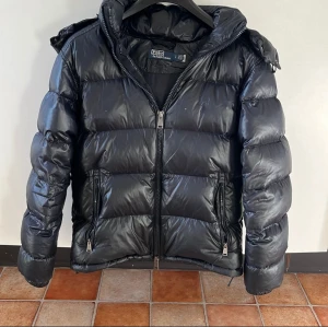 Svart pufferjacka från Polo Ralph Lauren - Säljer en svart(mörk blå)pufferjacka från Polo Ralph Lauren med huva och två dragkedjeförsedda fickor. Jackan har broderad logga på bröstet och en glansig finish. Perfekt för kalla dagar och riktigt snygg till streetwear-looken. Dm för snabb affär