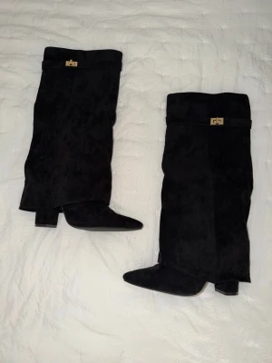 Svarta mockastövlar med klack - Snygga svarta stövlar i mjuk mocka med spetsig tå och klack. Stövlarna har en dekorativ guldfärgad detalj upptill och en stilren, slouchy passform. Perfekta för dig som gillar en klassisk men edgy look. Från Lululia.