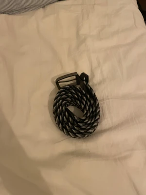 Svart och grå flätad tygbälte - Säljer ett snyggt flätat bälte i svart och grått med detaljer i läder vid ändarna och metallspänne. Bältet är i storlek 100 och har ett coolt mönster som passar till massor av jeans eller chinos. Perfekt accessoar för att lyfta din outfit. Hör av er vid frågor 🔥😁