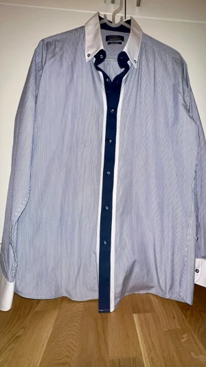 Snygg /Randig skjorta från Baurotti slim fit - i bra skick! - Snygg slim fit skjorta från Baurotti med tunna blå och vita ränder, vit krage och manschetter samt mörkblå knappslå. Långärmad modell i bomull med cool kontrast mellan detaljerna. Perfekt för dig som gillar stilrena och moderna skjortor.