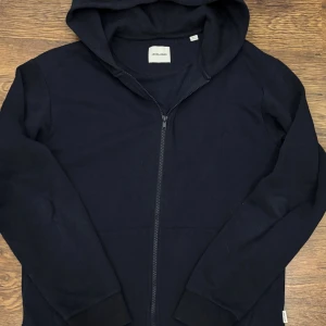 Mörkblå hoodie med dragkedja Jack & Jones - Snygg mörkblå hoodie från Jack & Jones med dragkedja framtill och huva. Tillverkad i mjukt sweat-material, perfekt för chill dagar. Klassisk design med ribbade muddar och diskret logga i nacken. Passar dig som gillar enkel och stilren streetwear. (Saknar snöre)