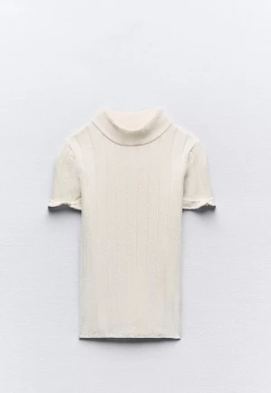 Ribbad polotopp i ljus beige - Säljer en stilren ribbad polotopp i ljus beige med korta ärmar och mjuk, stickad känsla. Toppen har en hög krage och subtila vertikala ränder som ger en clean look. Perfekt för dig som gillar minimalistisk stil och vill ha något som känns både chill och snyggt.