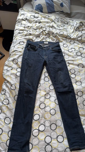 Tighta blå jeans från Tiger of Sweden - Snygga mörkblå jeans från Tiger of Sweden, modell Leon, med klassisk femficksdesign och kontrastsömmar. Jeansen har en rak och tight passform, tillverkade i slitstarkt denim och har en läderpatch med logga bak i midjan. Perfekt för dig som gillar stilrena och moderna jeans.