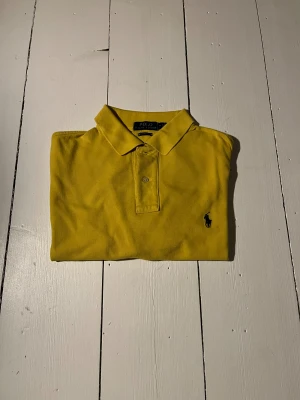 Gul piké från Polo Ralph Lauren - Klassisk gul pikétröja från Polo Ralph Lauren i modellen Classic Fit. Tröjan har krage, knappslå och den ikoniska broderade loggan på bröstet. Perfekt för en stilren och sportig look.