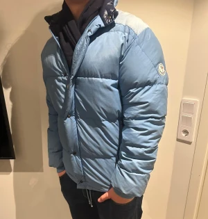 Ljusblå pufferjacka från Moncler - Säljer en ljusblå pufferjacka från Moncler med hög krage och dragkedja samt knappar framtill. Jackan har långa ärmar, två sidofickor med dragkedja och snörning nedtill. Materialet är polyester och dun, perfekt för kalla dagar. Moncler-logga på ärmen.