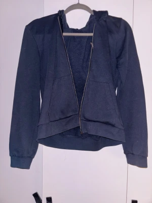 Mörkblå hoodie med dragkedja - Säljer en mörkblå hoodie med huva och praktisk dragkedja framtill. Tröjan har långa ärmar, ribbade muddar och två stora fickor på magen. Perfekt för chill dagar eller när du vill ha något bekvämt och enkelt.