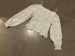 Blommig blus med smock och volang - Supersöt blus med ljusblått och vitt blommönster, smockad överdel och volangkant nertill. Långa puffiga ärmar med resår vid handlederna och tunna axelband. Perfekt för en romantisk look och passar till jeans eller kjol.