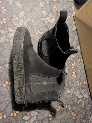 Svarta Chelsea boots från Canada Snow - Säljer ett par svarta Chelsea boots från Canada Snow med grov platt sula och dragflikar både fram och bak. Skorna är tillverkade i mocka med detaljer i skinn och har en stilren, rund tå. Perfekta för höst och vinter med sin robusta design.