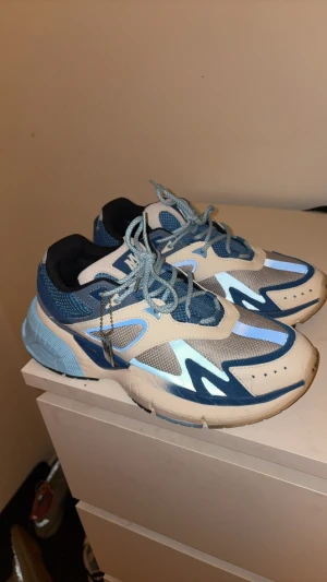 AMIRI blå och vita sneakers strl 44 - Säljer ett par AMIRI sneakers i storlek 44 med snygg design i olika blå toner, vitt och grått. Skorna har mesh och skinnpaneler, ljusblå snörning och AMIRI-logga på hälen. Platt sula och rund tå, perfekt för dig som gillar streetwear och exklusiva märken.