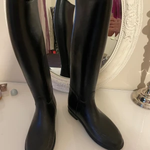 Svarta höga ridstövlar i gummi - Säljerw ett par svarta höga ridstövlar i gummi, klassisk modell med platt sula och rund tå. Stövlarna är tillverkade i Israel och har en stilren, minimalistisk look som passar perfekt till ridning. Storlek 36, med diskret präglad märkning under sulan.