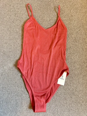 Svart baddräkt black bathingsuit becksöndergaard - En fantastiskt fin och feminin baddräkt från Becksöndergaard, helt ny med tags och aldrig använd. Baddräkten kommer i en mjuk, varm rosaton med ett vackert subtilt glittrande ribbat tyg som fångar ljuset och ger en exklusiv känsla.  Modellen har en smickrande V-ringning framtill och tunna, justerbara axelband för perfekt passform. Baksidan har en något djupare skärning för ett stilrent och elegant uttryck. Materialet är både stretchigt och bekvämt med en helfodrad insida som ger stöd och komfort