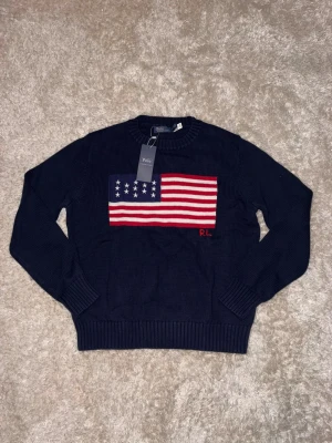 Mörkblå stickad tröja Polo Ralph Lauren - Snygg mörkblå stickad tröja från Polo Ralph Lauren med amerikansk flagga i rött, vitt och blått på bröstet samt RL-broderi. Rund hals och ribbade muddar vid ärmslut och nederkant. Perfekt för dig som gillar klassisk stil med en twist.