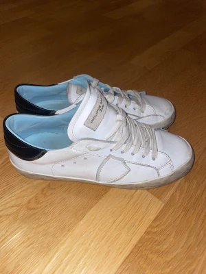 Philippe Model vita sneakers med svart häl - OBS! Låda kommer med skorna. Snygga vita sneakers från Philippe Model med svart detalj på hälen och ljusblå insida. Skorna har snörning, rund tå och platt sula. Tillverkade i skinn med diskreta grå detaljer och logga på tungan. Perfekta för dig som gillar stilrena och klassiska sneakers.