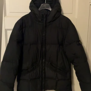 Svart pufferjacka från Stone Island Junior - Säljer en svart pufferjacka från Stone Island med huva och dragkedja framtill. Jackan har den klassiska loggan på ärmen och två snedställda fickor framtill. Riktigt varm och skön. Passar allt ifrån 160 till 180, jag är 190 och den passar nästan mig, lite kort. Skriv för mer bilder. Ny pris ca 7000 kr. Finns 2 st små hål som syns väldigt väldigt lite. Köpt från flannels.com för cirka 5 år sedan 