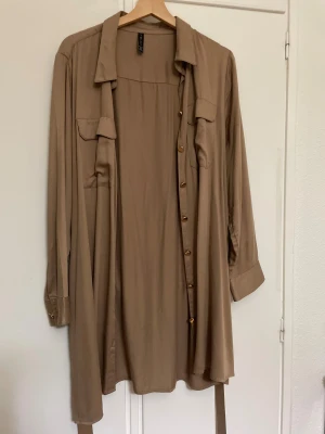 Beige skjortklänning från Amisu XL - Stilren beige skjortklänning från Amisu i storlek XL. Klänningen har långa ärmar, knappar framtill, två bröstfickor och ett bälte i midjan för snygg passform. Perfekt för dig som gillar en avslappnad men trendig look.