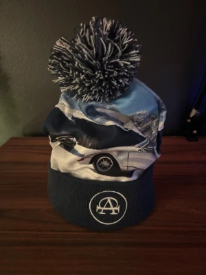 Mössa med bilmotiv och tofs med märket all cap one! - Snygg mössa med stor svartvit tofs och tryck av klassiska sportbilar mot ett vinterlandskap. Nedre delen är mörkblå med broderad All cap one logga. Perfekt för dig som gillar bilar och vill sticka ut i vinter. Materialet är mjukt och värmande tyg.