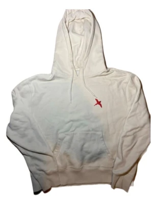 Vit hoodie med röd fågelbrodyr - Vit hoodie med huva och dragsko, samt en broderad röd fågel på bröstet. Klassisk känguruficka framtill och ribbade muddar. Perfekt för dig som gillar stilrena plagg med en liten detalj som sticker ut.