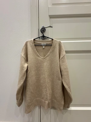 Beige stickad v-ringad tröja H&M - Mysig beige stickad tröja från H&M med v-ringad hals och långa ärmar. Tröjan har ribbade muddar vid ärmslut och nederkant, vilket ger en chill och avslappnad vibe. Perfekt för lager-på-lager under hösten och vintern.