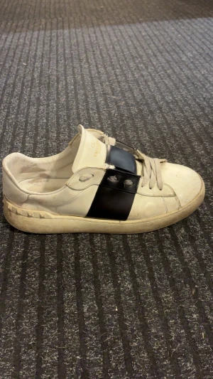 Valentino open - Snygga Valentino Garavani Open sneakers i vitt skinn med en bred svart panel över mitten. Klassisk rund tå, platt sula och grå snörning. Diskret nitar på hälen och logga på plösen. Perfekt för dig som gillar statement-detaljer och lyxig streetstil. Skriv om ni har frågor eller funderingar!😊