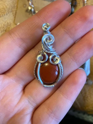 Handgjord wire wrap-hänge med röd jaspis - Unikt hänge med en rund, brunröd sten infattad i detaljerad silverfärgad wire wrap-design. Spiralmönster och tvinnad metall ger en bohemisk och konstnärlig vibe. Perfekt som statement piece till kedja eller läderband.