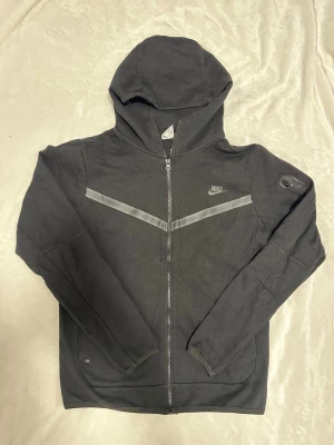Svart Nike tech hoodie med dragkedja S - Svart hoodie från Nike i storlek S med hel dragkedja, huva och två fickor. Snyggt grått V-mönster över bröstet och Nike-logga på vänster sida. Materialet är mjukt och bekvämt, perfekt för chill eller träning. Extra ficka med dragkedja på ärmen.