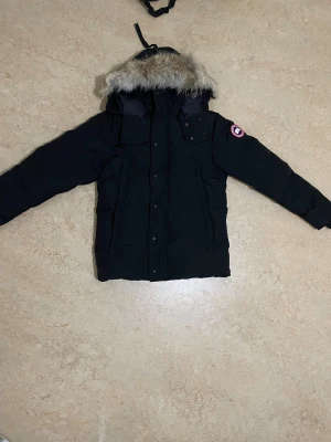 Svart dunjacka Canada Goose med päls - Säljer en svart dunjacka från Canada Goose med avtagbar huva och pälsdetalj. Jackan har klassisk patch på ärmen, dragkedja och knappar framtill samt flera fickor. Perfekt för kalla vinterdagar och riktigt snygg streetstil.