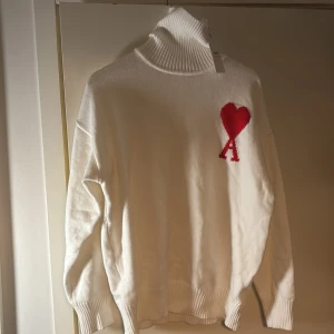 Vit polotröja från Ami med rött hjärta - Vit polotröja från Ami Alexandre Mattiussi med ribbad hög krage och muddar. Tröjan har ett rött hjärta med bokstaven A broderat på bröstet. Oversized passform och stickad i mjukt material, perfekt för kyliga dagar.