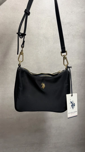 Svart axelväska från U.S. Polo Assn. - Stilren svart axelväska från U.S. Polo Assn. med justerbar rem och guldfärgade metalldetaljer. Väskan har dragkedja upptill och en liten logga framtill. Perfekt storlek för det viktigaste och enkel att matcha till olika outfits.