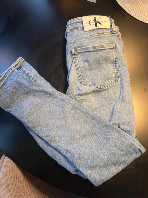 Ljusblå jeans från Calvin Klein - Snygga ljusblå jeans från Calvin Klein Jeans med klassisk femficksdesign och tydlig CK-logga på bakfickan. Jeansen har rak passform och är tillverkade i mjukt denimtyg som ger en skön känsla. Perfekt för dig som gillar stilrena och tidlösa jeans.