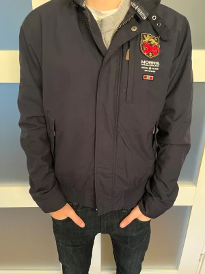 Morris marinblå vindjacka  - Snygg marinblå vindjacka från Morris med broderad logga och emblem på bröstet. Jackan har ståkrage med tryckknapp, dragkedja och ribbade muddar. Fodret har röd och vit detalj. Perfekt för dig som gillar klassisk stil med coola detaljer.