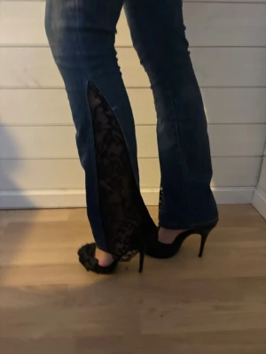 Blå bootcut jeans med spetsdetalj - Unika blå jeans med bootcut passform och svarta spetsinsatser längs bensluten. Jeansen har klassisk femficksdesign, låg midja och coola slitningar framtill. Perfekt för dig som vill sticka ut med en edgy och trendig look. Jeansen är originellt från Crocker Jean, men det är vi på TÄFmade som sytt om dom! Jeansen är i storlek 25 längd 30! 